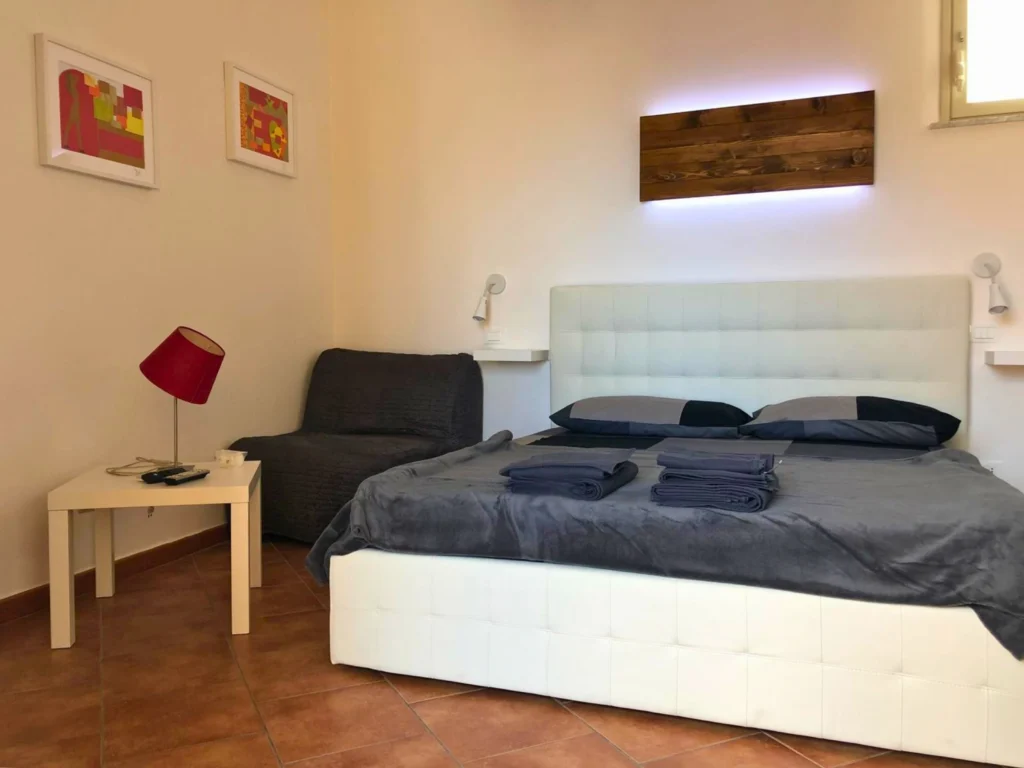 Camera-da-letto-moderna-e-minimalista-con-letto-in-pelle-bianca-e-testata-tufted-biancheria-da-letto-grigia-scura-due-asciugamani-grigi-sul-letto-mensole-bianche-con-lampade-da-lettura-parete-con-illuminazione-a-pannelli-di-legno-due-quadri-astratti-con-toni-rossi-gialli-e-arancioni