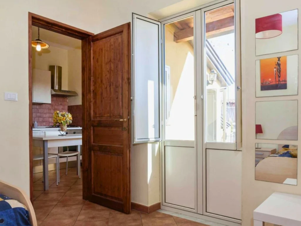 Vista-interni-con-porta-di-legno-aperta-su-cucina-tavolo-bianco-con-fiori-e-sedie-aspiratore-e-armadietti-in-cucina-a-destra-ampia-finestra-su-area-esterna-parete-con-tre-quadri-persona-su-sfondo-arancione-letto-con-coperta-a-scuri-e-gialli-a-sinistra