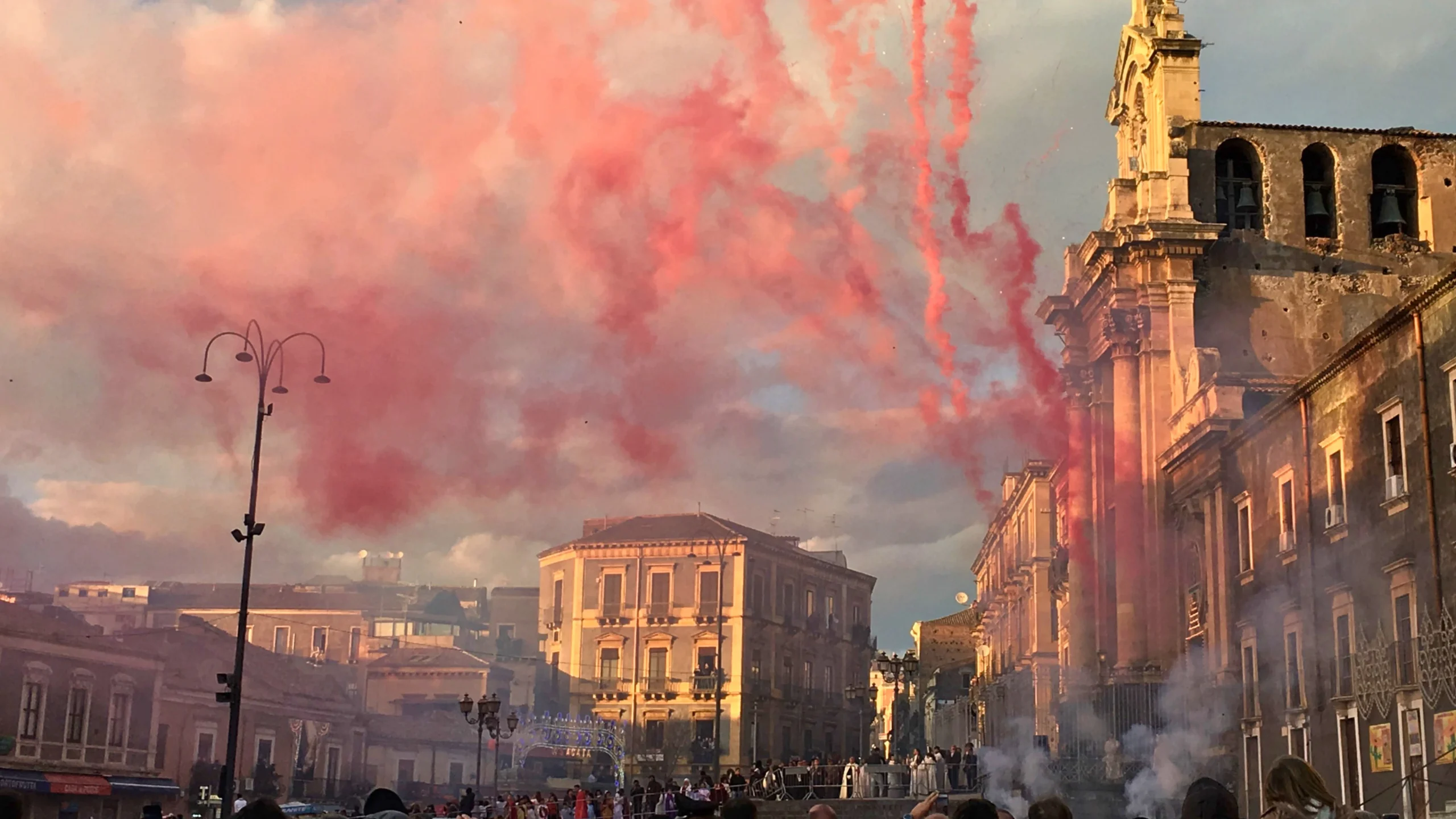 Grande-folla-radunata-nella-storica-Piazza-Carlo-Alberto-a-Catania-durante-la-Festa-di-Sant'Agata-con-edifici-antichi-e-una-chiesa-sullo-sfondo-fumo-rosso-o-fuochi-d'artificio-nel-cielo-celebrazione-vibrante-e-colorata