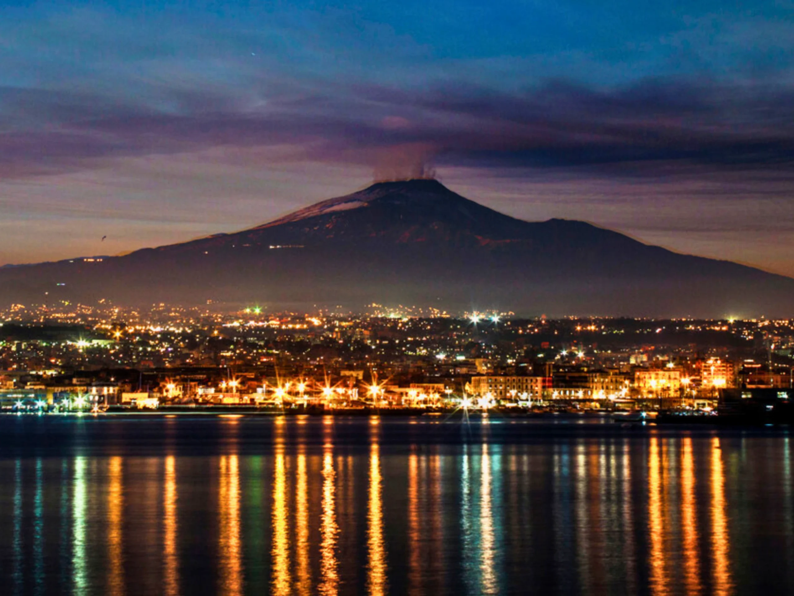 Veduta-notturna-di-Catania-con-luci-della-città-riflesse-nel-mare-e-Monte-Etna-sullo-sfondo-con-una-leggera-nuvola-di-fumo-in-cima-cielo-scuro-con-gradiente-di-colori-dal-blu-al-viola-suggestiva-immagine-di-città-e-vulcano