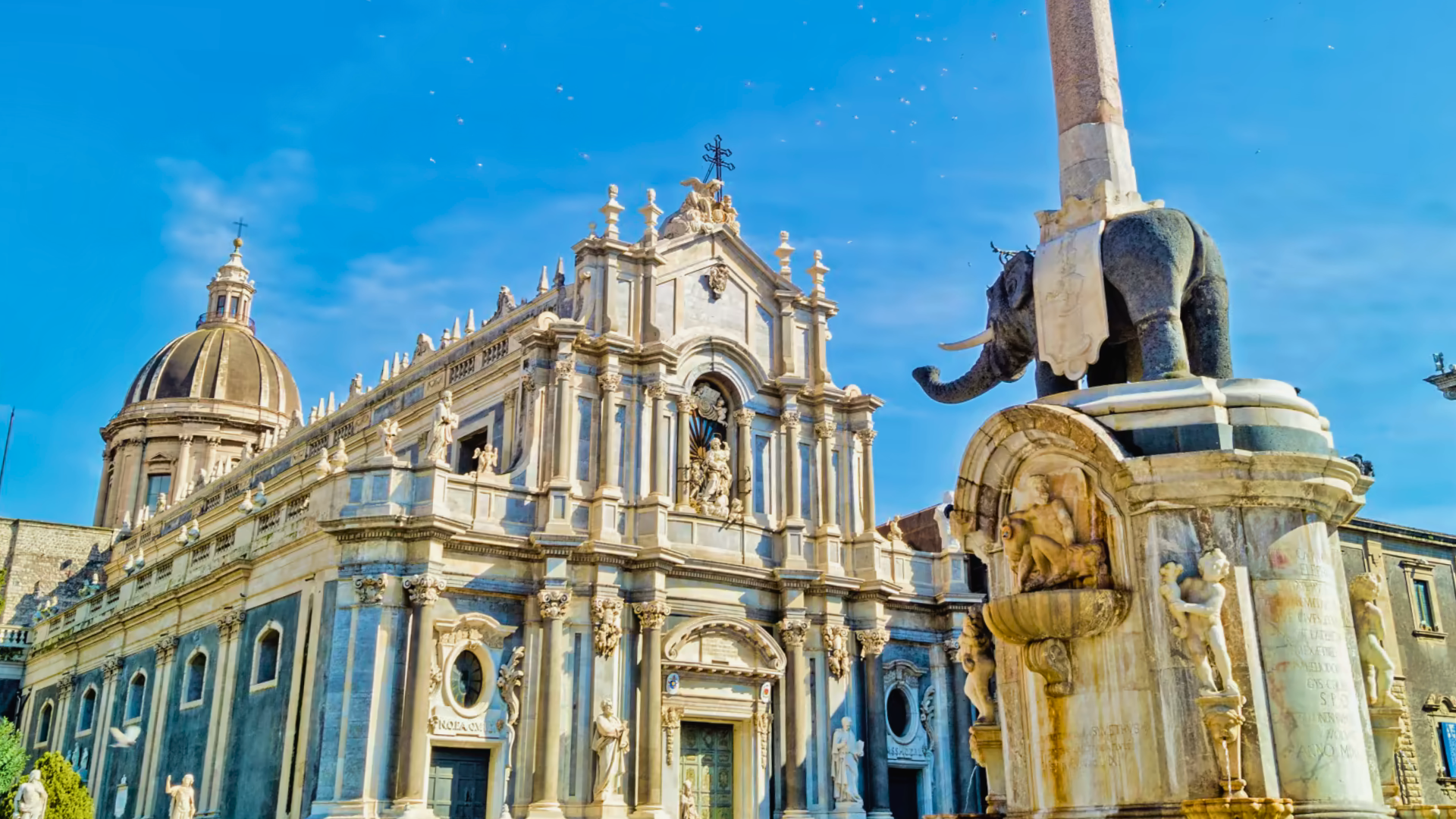 Piazza-Duomo-di-Catania-con-la-facciata-storica-del-Duomo-dalla-dome-grigia-e-numerose-statue-statua-di-un-elefante-che-porta-un-obelisco-sulla-destra-cielo-azzurro-limpido-con-edifici-decorativi-sullo-sfondo