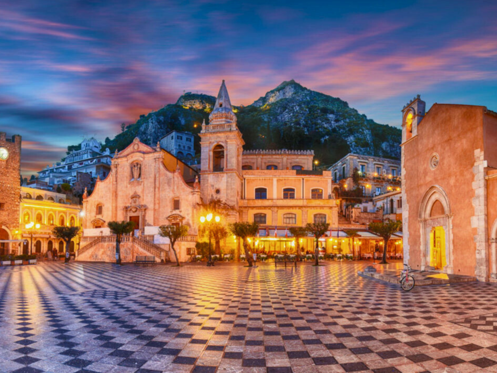 Piazza-IX-Aprile-a-Taormina-con-pavimentazione-a-scacchi-bianca-e-nera-torre-dell'orologio-a-sinistra-chiesa-con-campanile-al-centro-edifici-storici-con-balconi-archi-e-decorazioni-collina-con-edifici-e-verde-sullo-sfondo-cielo-al-tramonto