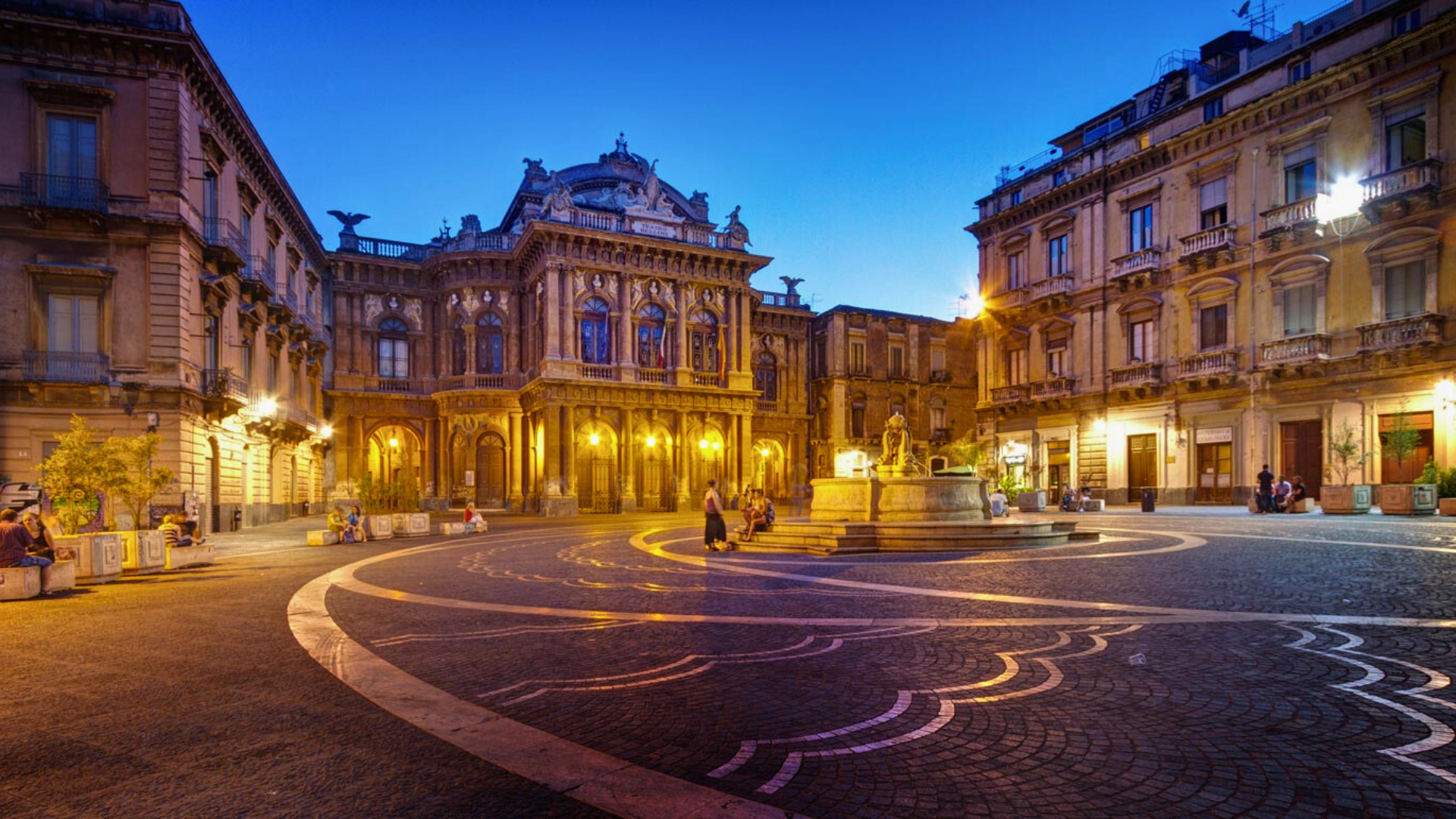 casa-vacanze-in-sicilia-vicino-Piazza-Teatro-Massimo-di-Catania-Italia-con-il-grande-Teatro-Massimo-Bellini-riccamente-decorato-in-stile-Barocco-e-Rinascimentale-facciata-intricata-con-statue-e-finestre-ad-arco-pavimentazione-decorativa-fontana-centrale-con-statua-illuminazione-calda-e-persone-che-camminano