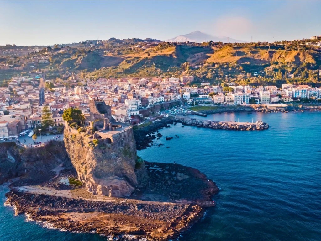 Vista-aerea-di-Aci-Castello-Sicilia-con-il-Norman-Castle-di-Aci-su-una-roccia-sul-mare-edifici-della-città-densamente-disposti-con-architettura-tradizionale-e-moderna-piccolo-porto-con-molo-colline-sullo-sfondo-e-Monte-Etna-illuminati-dalla-luce-del-tardo-pomeriggio