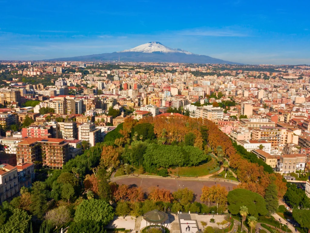 Veduta-aerea-della-città-di-Catania-con-il-vulcano-Etna-innevato-sullo-sfondo-grande-parco-verde-in-primo-piano-edifici-residenziali-e-commerciali-suggestivo-contrasto-tra-urbanizzazione-e-natura