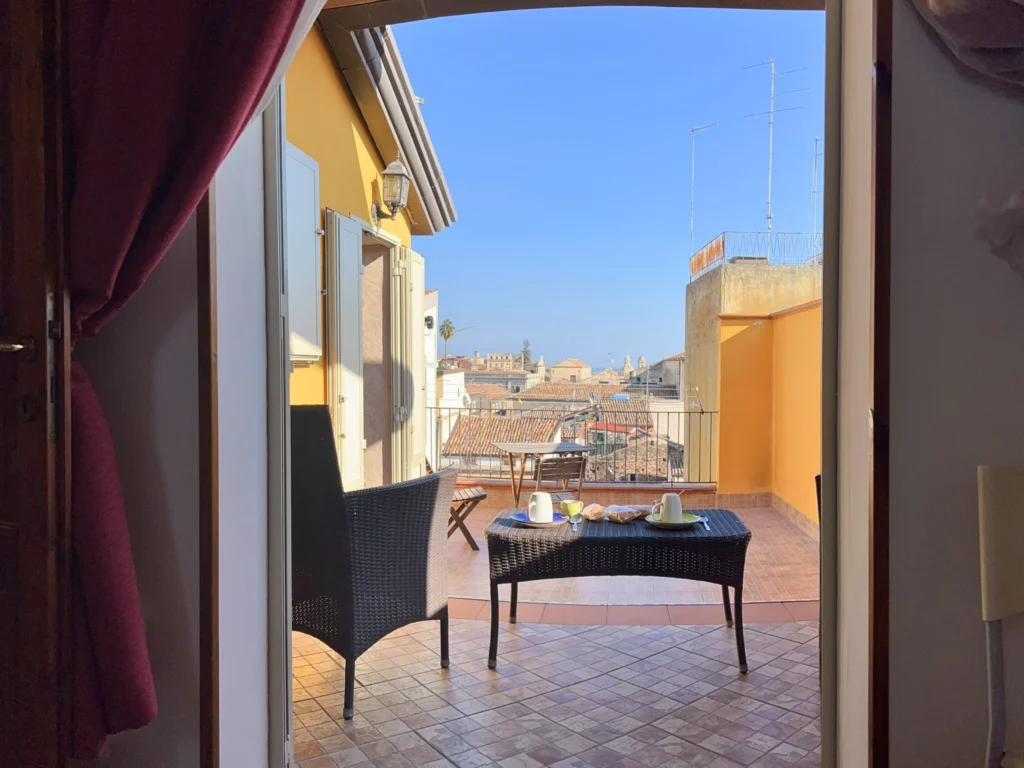 Vista-dall'interno-di-una-stanza-dell-attico-a-catania-su-una-terrazza-con-pavimento-piastrellato-sedie-in-vimini-nera-e-tavolo-nero-con-teiera-e-due-tazze-sullo-sfondo-edifici-con-tetti-in-terracotta-e-cielo-azzurro-incorniciato-da-porta-aperta-con-tende-rosse