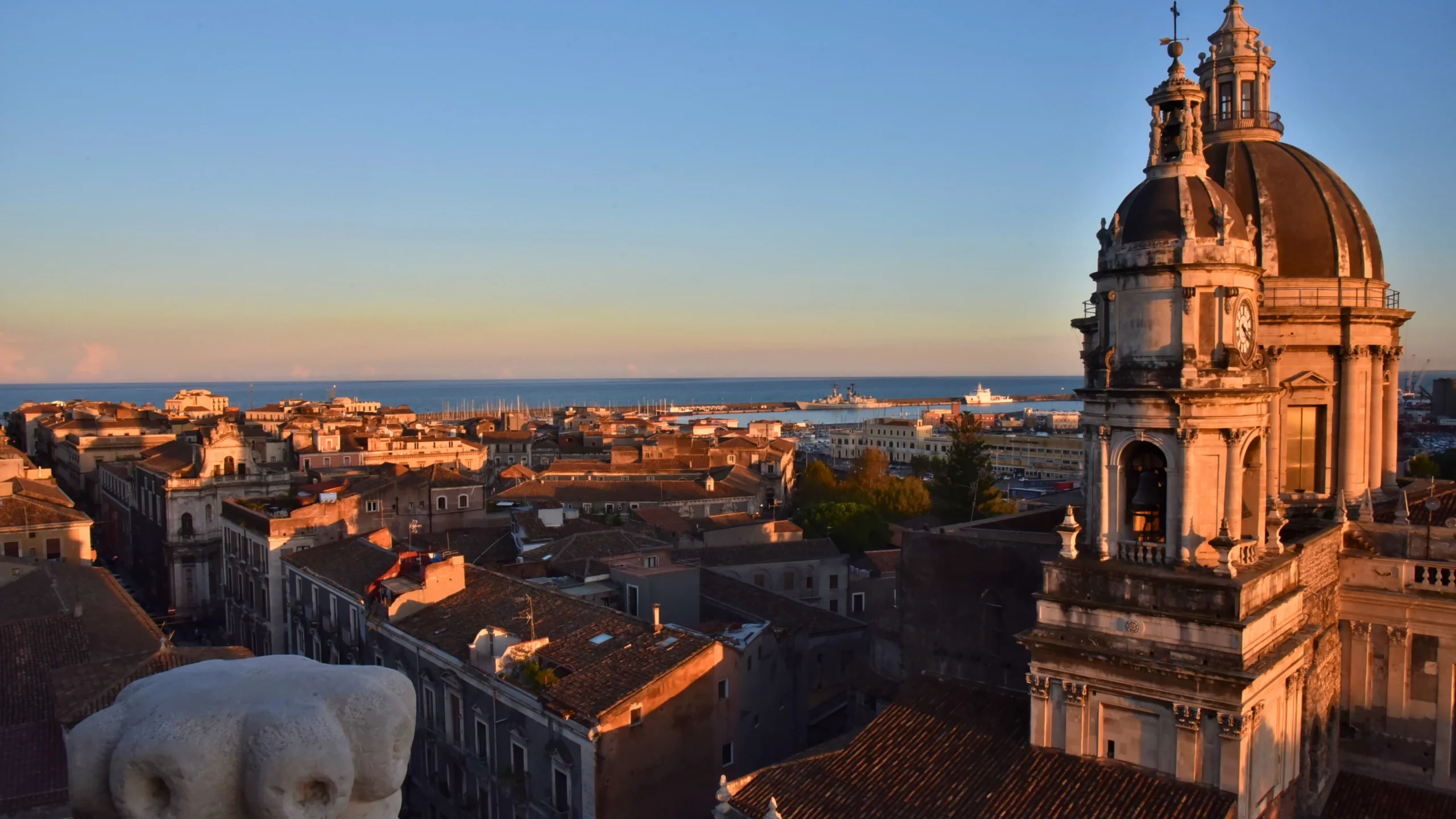 Panoramica-di-Catania-al-tramonto-con-cupola-del-Duomo-in-primo-piano-area-urbana-densa-sullo-sfondo-porto-con-barche-cielo-chiaro-con-tonalità-dorate