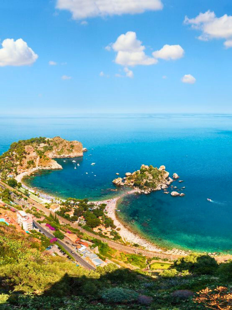 Vista-panoramica-dell'Isola-Bella-a-Taormina-con-coste-rocciose-acque-blu-cristalline-vegetazione-rigogliosa-e-una-piccola-isola-collegata-alla-terraferma-da-una-stretta-striscia-di-terra-collina-densa-di-edifici-verdeggianti-sullo-sfondo-e-cielo-azzurro-con-nuvole-sparse