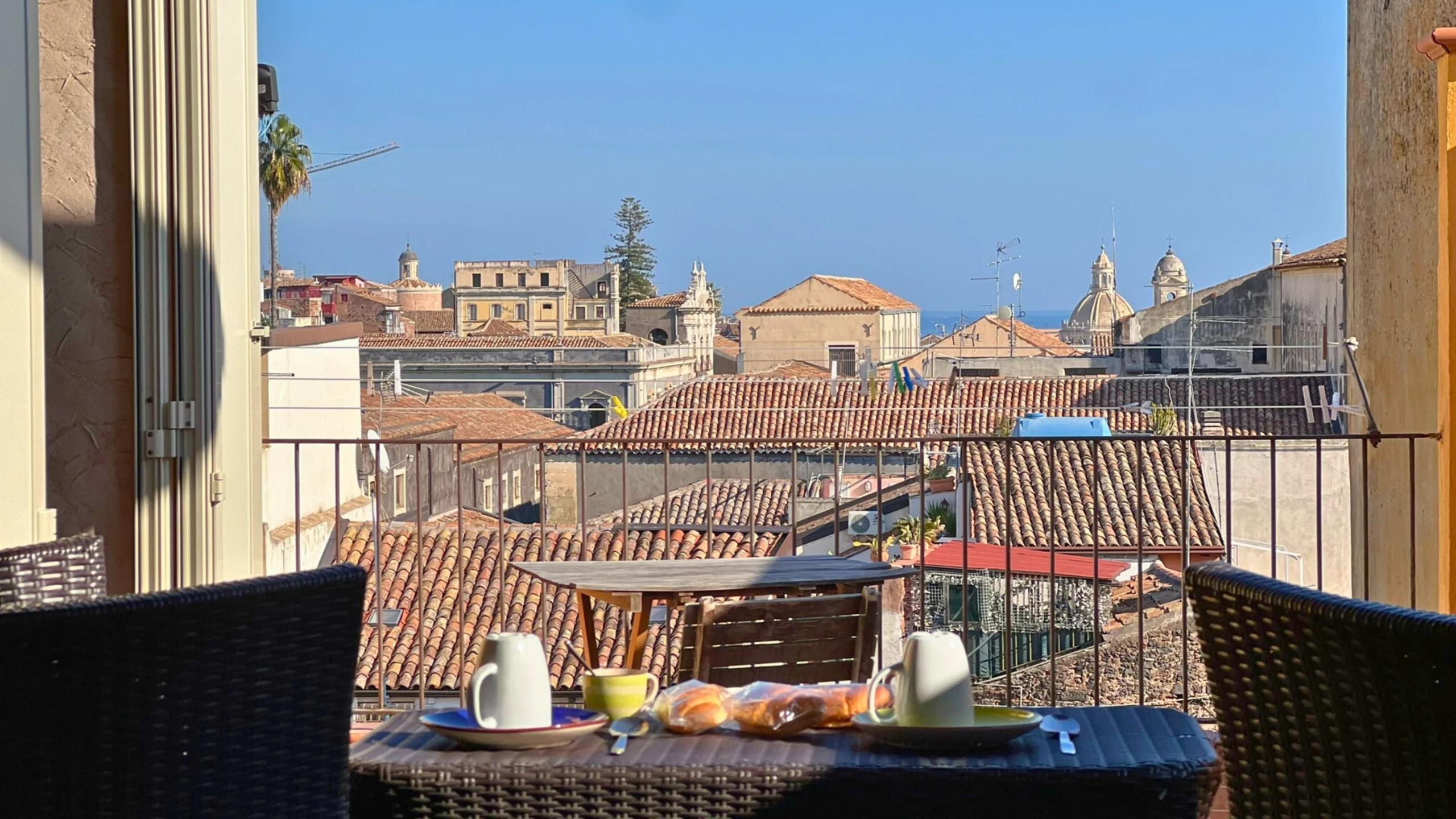 Vista-dal-balcone-dell'attico-su-Catania-tavolo-per-colazione-con-teiera-tazze-e-dolci-sullo-sfondo-si-vedono-edifici-storici-e-la-cupola-del-duomo-di-Catania