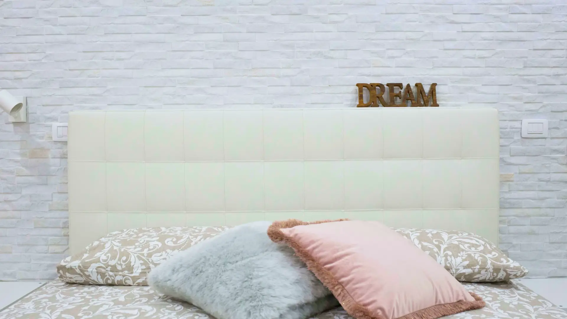 Letto-con-testata-in-pelle-bianca-contro-parete-di-mattoni-bianchi-coperta-decorata-in-beige-e-bianco-con-cuscini-grigi-e-rosa-interno-moderno-e-accogliente-con-illuminazione-murale-e-segnale-di-legno-DREAM