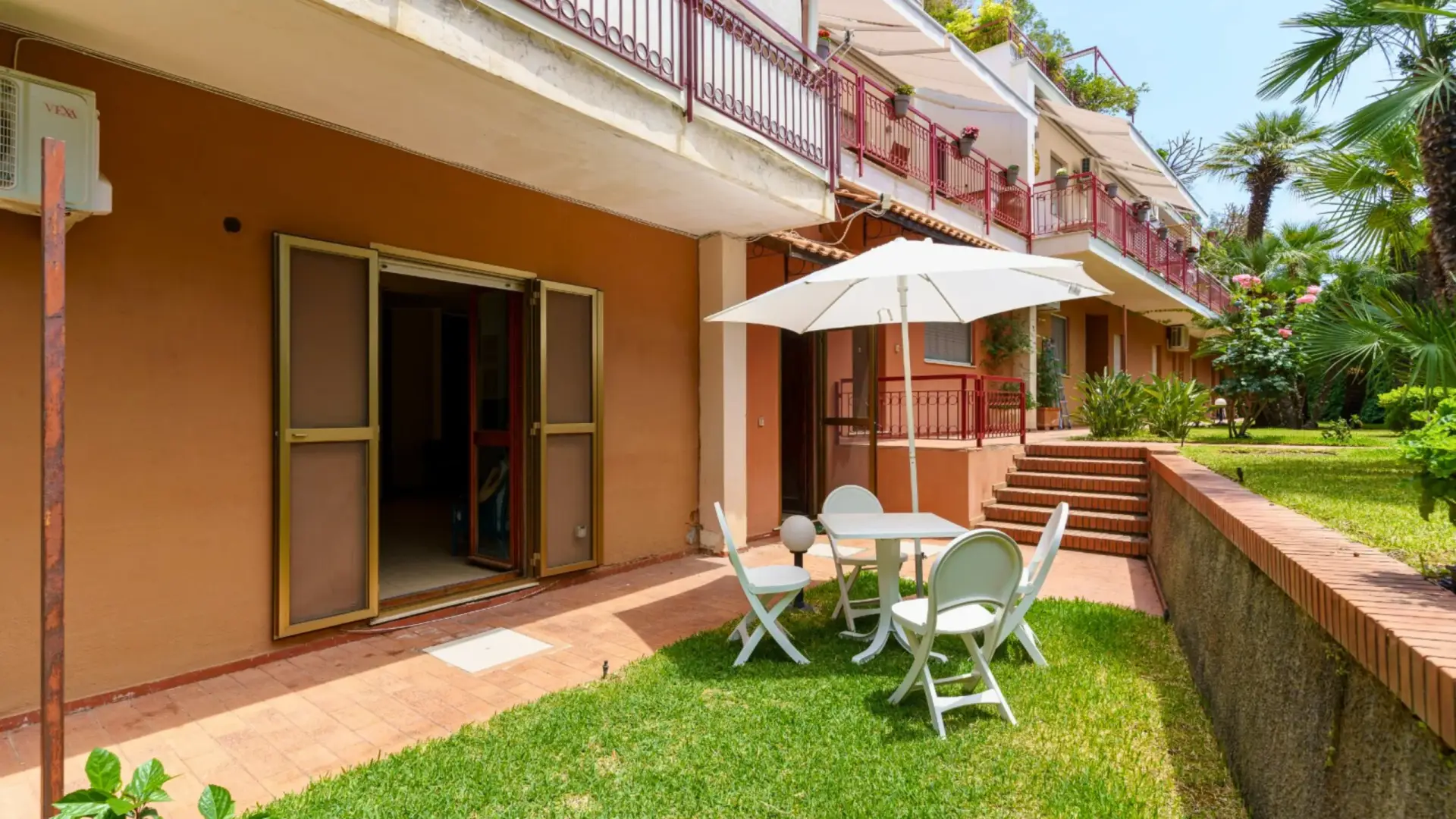 Giardino-privato-di-una-residenza-con-patio-attrezzato-tavolo-bianco-con-quattro-sedie-e-grande-ombrello-bianco-sul-tavolo-edificio-con-esterno-rossiccio-e-balcone-verde-lussureggiante-con-palme-e-piante-fiorite-scale-che-portano-ad-un-livello-superiore-ambiente-tranquillo-e-ben-curato