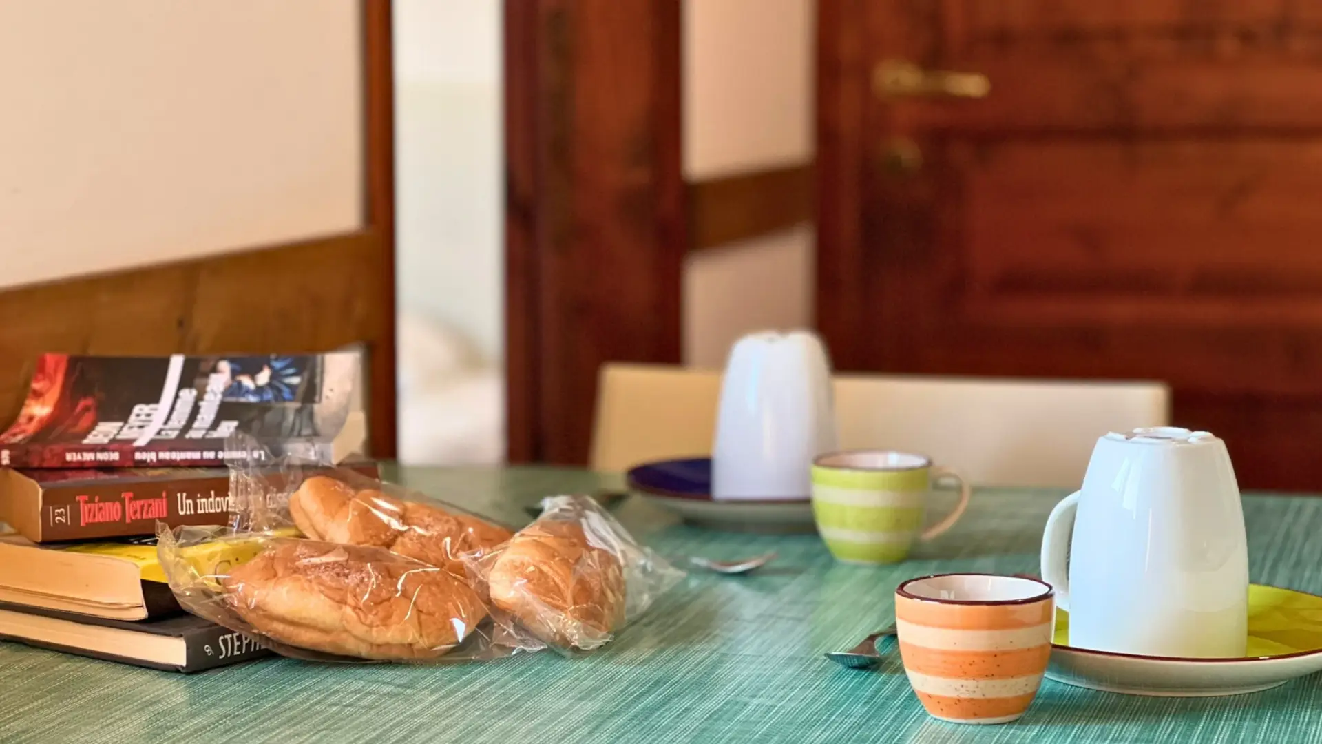 Tavola-da-colazione-con-due-piatti-e-tazze-capovolte-tazza-a-strisce-verdi-e-tazza-a-strisce-arancioni-cucchiai-vicino-ai-piatti-sacchetto-di-plastica-con-croissant-libri-impilati-sul-tavolo-sfondo-con-porta-di-legno-e-parete-ambiente-accogliente