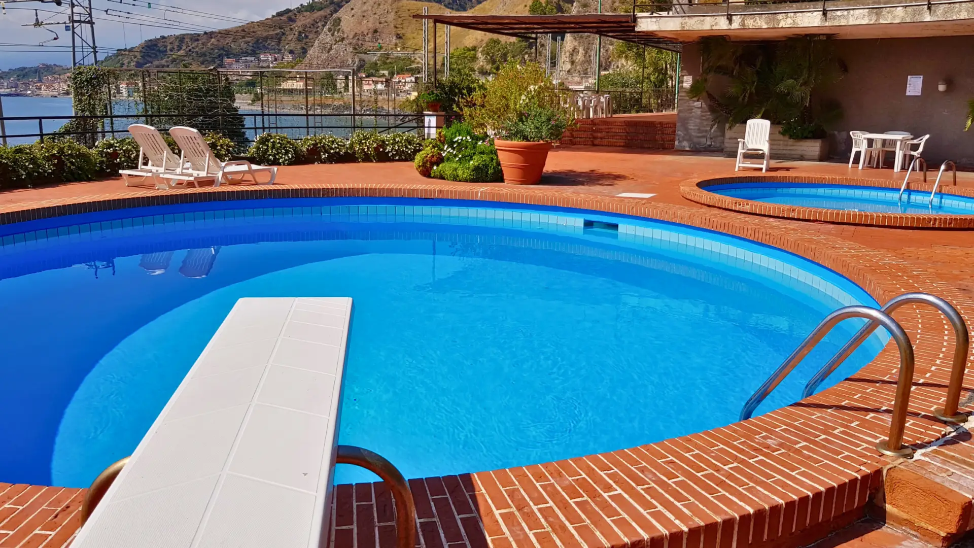 Vista-delle-piscine-del-residence-Capo-Taormina-House-una-piscina-per-adulti-con-trampolino-e-una-piscina-piccola-per-bambini-area-pavimentata-con-mattoni-rossi-sdraio-e-tavoli-con-sedie-vegetazione-e-piante-in-vasi-sullo-sfondo-paesaggio-montuoso-e-corpo-d'acqua-mare