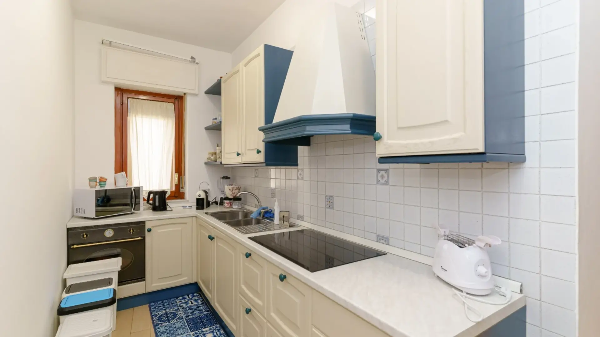 Cucina-moderna-con-armadietti-bianchi-e-blu-paraschizzi-di-piastrelle-bianche-con-decorazioni-piccole-rubinetto-e-lavello-di-acciaio-inossidabile-elettrodomestici-da-cucina-come-microonde-bollitore-macchina-da-caffè-e-tostapane-sul-top-dei-mobili-tappeto-pavimento-con-motivo-blu-e-bianco-finestra-con-tenda-ambiente-ordinato-con-mix-di-elementi-tradizionali-e-contemporanei