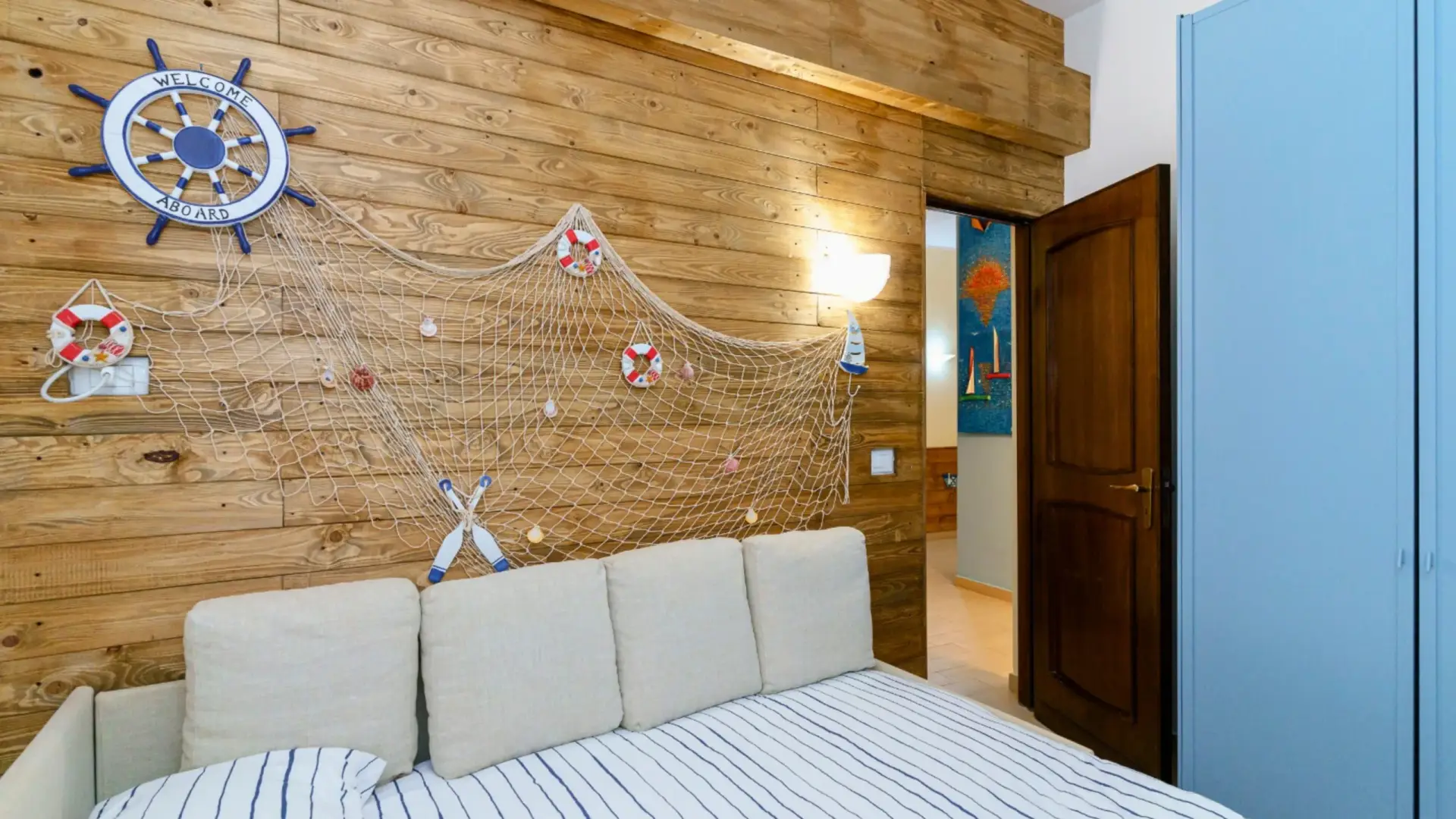 Camera-da-letto-al-Capo-Taormina-House-con-tema-nautico-parete-in-legno-con-rete-da-pesca-piccoli-salvagente-timone-navale-con-scritta-WELCOME-ABOARD-letto-con-coperta-a-righe-e-cuscini-beige-porta-di-legno-aperta-contro-parete-pittura-colorata-armadio-blu-luce-naturale