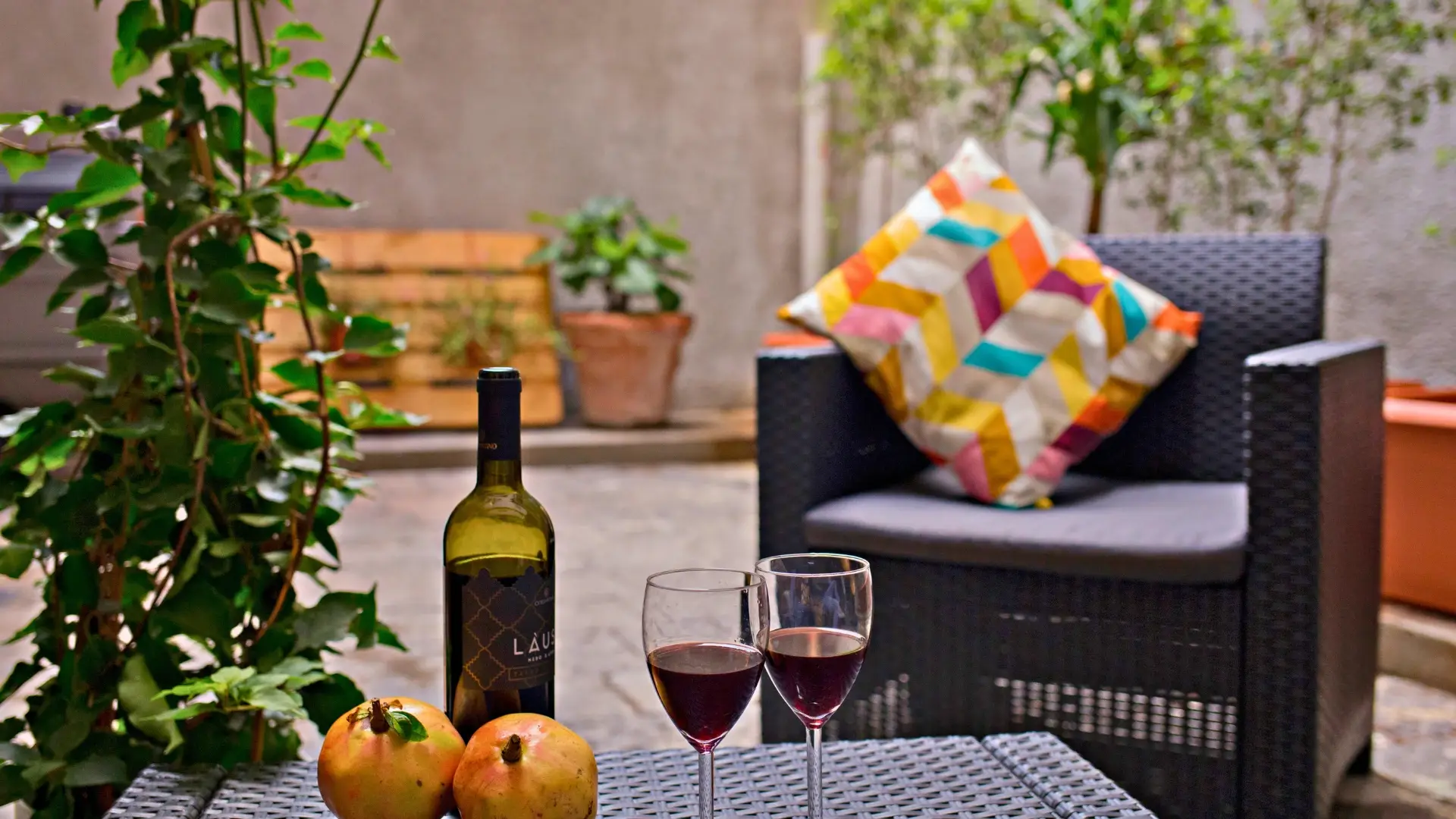 Cortile-esterno-con-tavolo-di-vimini-bottiglia-di-vino-rosso-due-bicchieri-di-vino-due-melograni-sedia-in-vimini-con-cuscino-geometrico-colorato-piante-in-vaso-e-fioriera-di-legno-in-sfondo