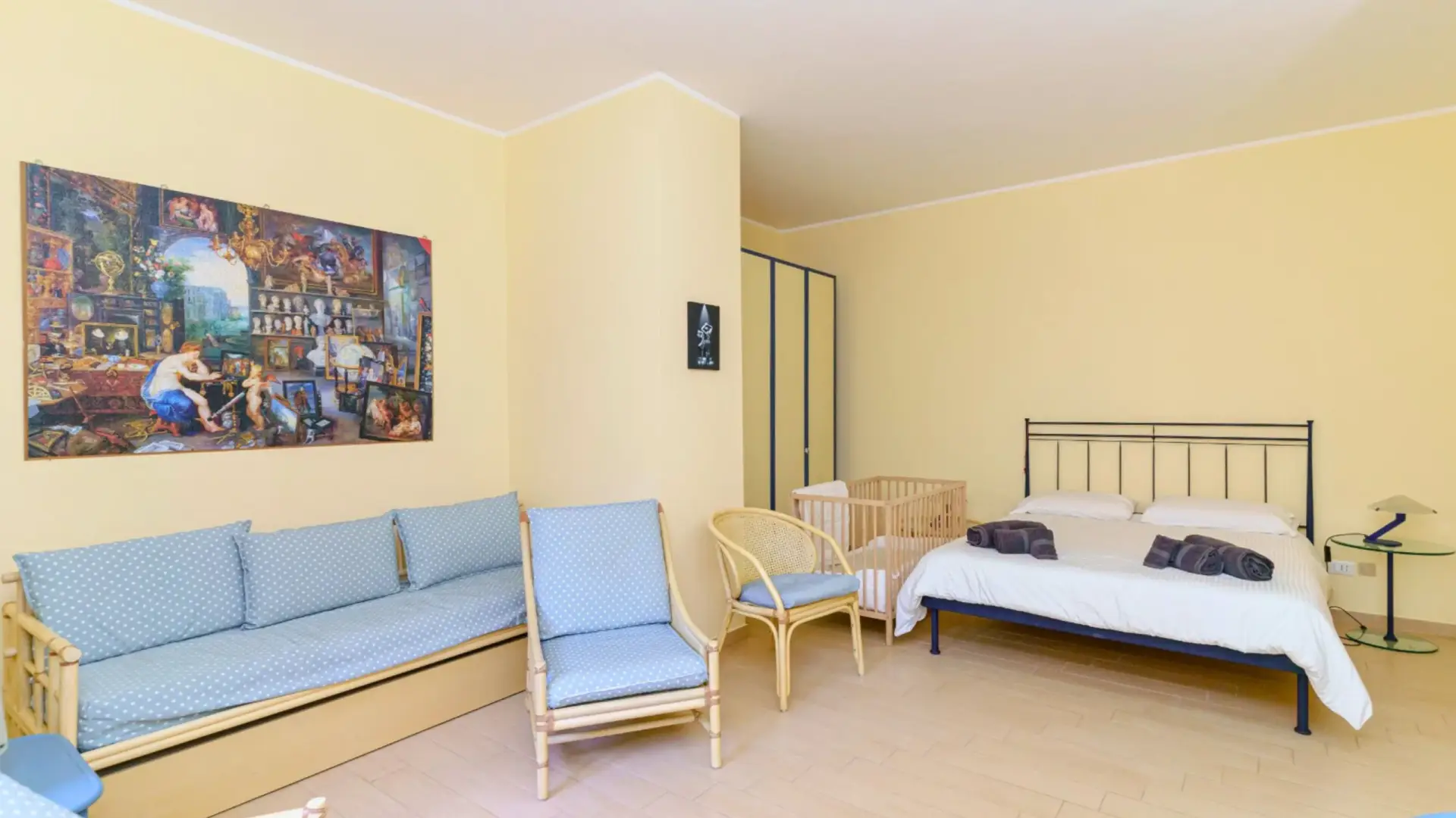 Vista-della-camera-da-letto-principale-nel-Capo-Taormina-House-stanza-spaziosa-con-pareti-chiare-letto-matrimoniale-con-lenzuola-bianche-e-asciugamani-piegati-divano-con-cuscini-blu-a-pois-bianchi-due-sedie-coordate-lettino-per-bambini-armadio-con-ante-scorrevoli-e-tavolino-con-lampada-grande-quadro-artistico-appeso-alla-parete-atmosfera-luminosa-e-accogliente